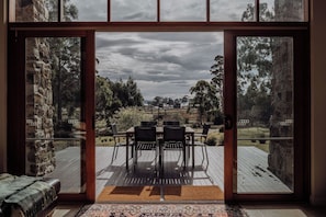 A view through a glass door to an outdoor deck with a table and chairs, surrounded by stone walls and trees.