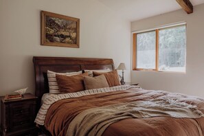 A bedroom with a wooden headboard, a bedside table with books, a lamp, and a window with a view of trees. - Adventure Bay Retreat (Adventure Bay)