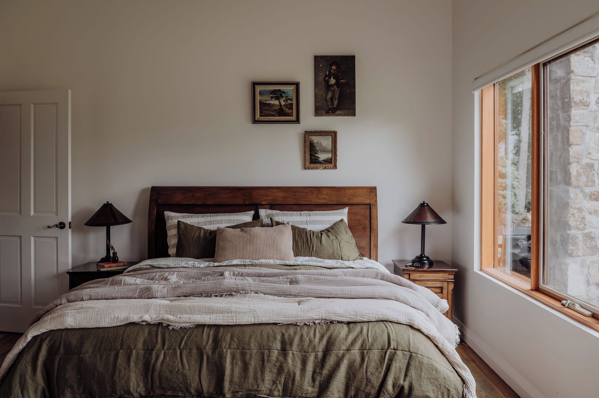 Une chambre à coucher avec une tête de lit en bois, un lit recouvert d’une couverture à rayures, deux tables de chevet avec des lampes et trois tableaux encadrés accrochés au mur.