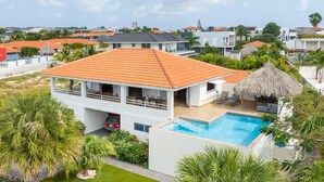 A modern two-story house with a swimming pool, surrounded by lush greenery and palm trees.