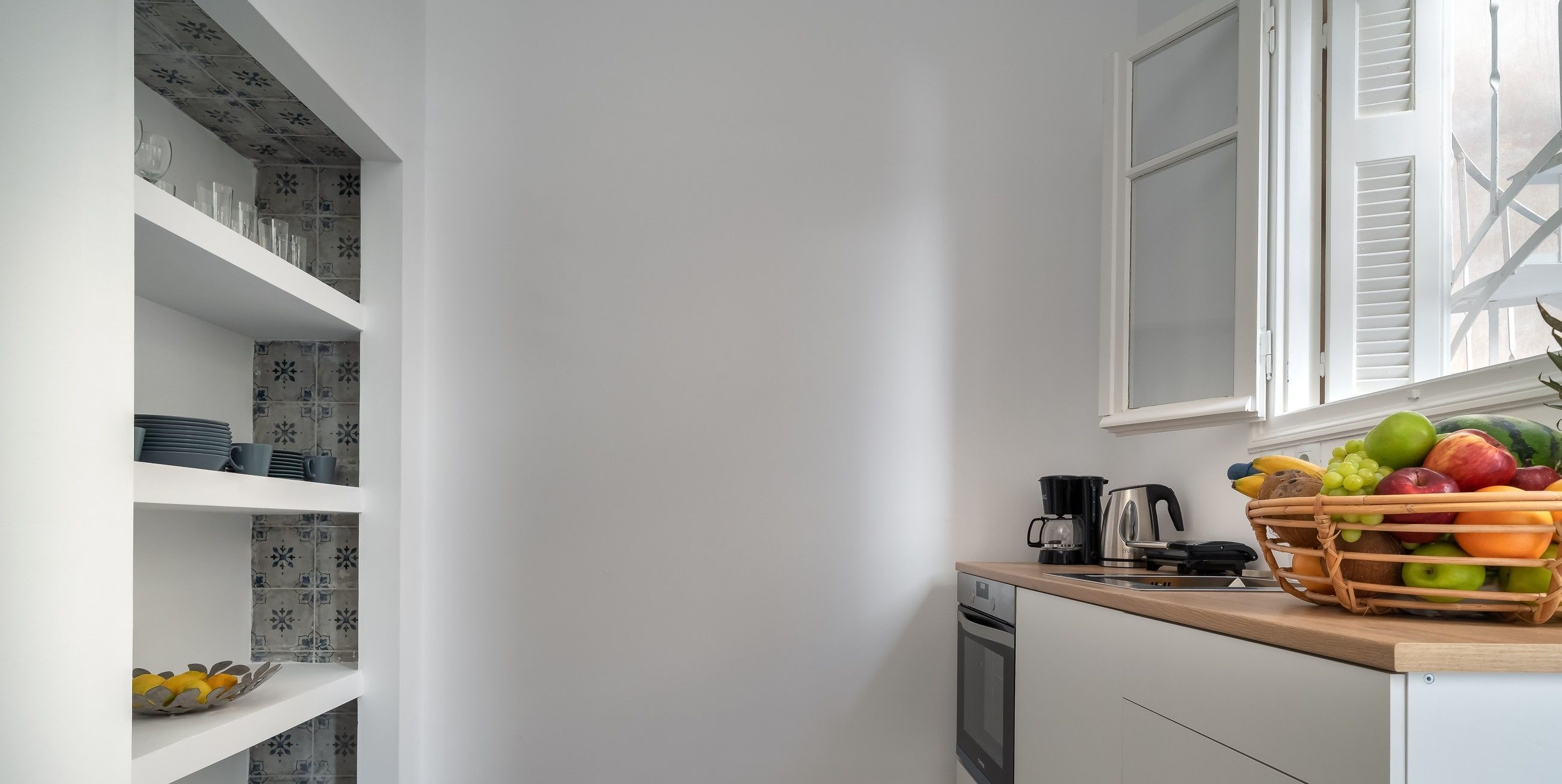 A kitchen with white walls, a wooden countertop, and a window with shutters. There is a basket of fruit on the counter and shelves with dishes and a decorative bowl.