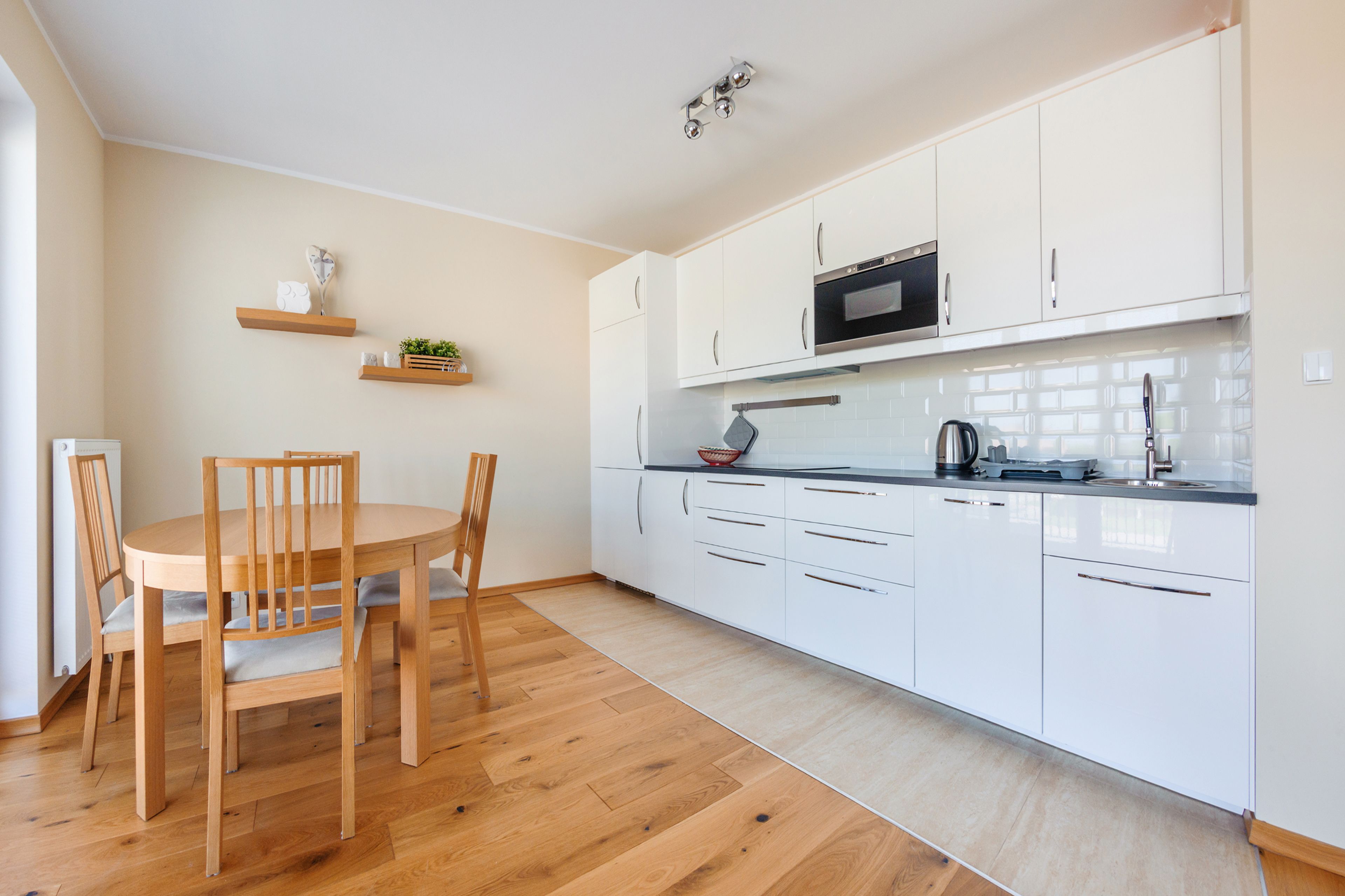 A modern kitchen with white cabinets, a microwave, and a sink. There is a dining table with chairs and a small shelf with decorative items.