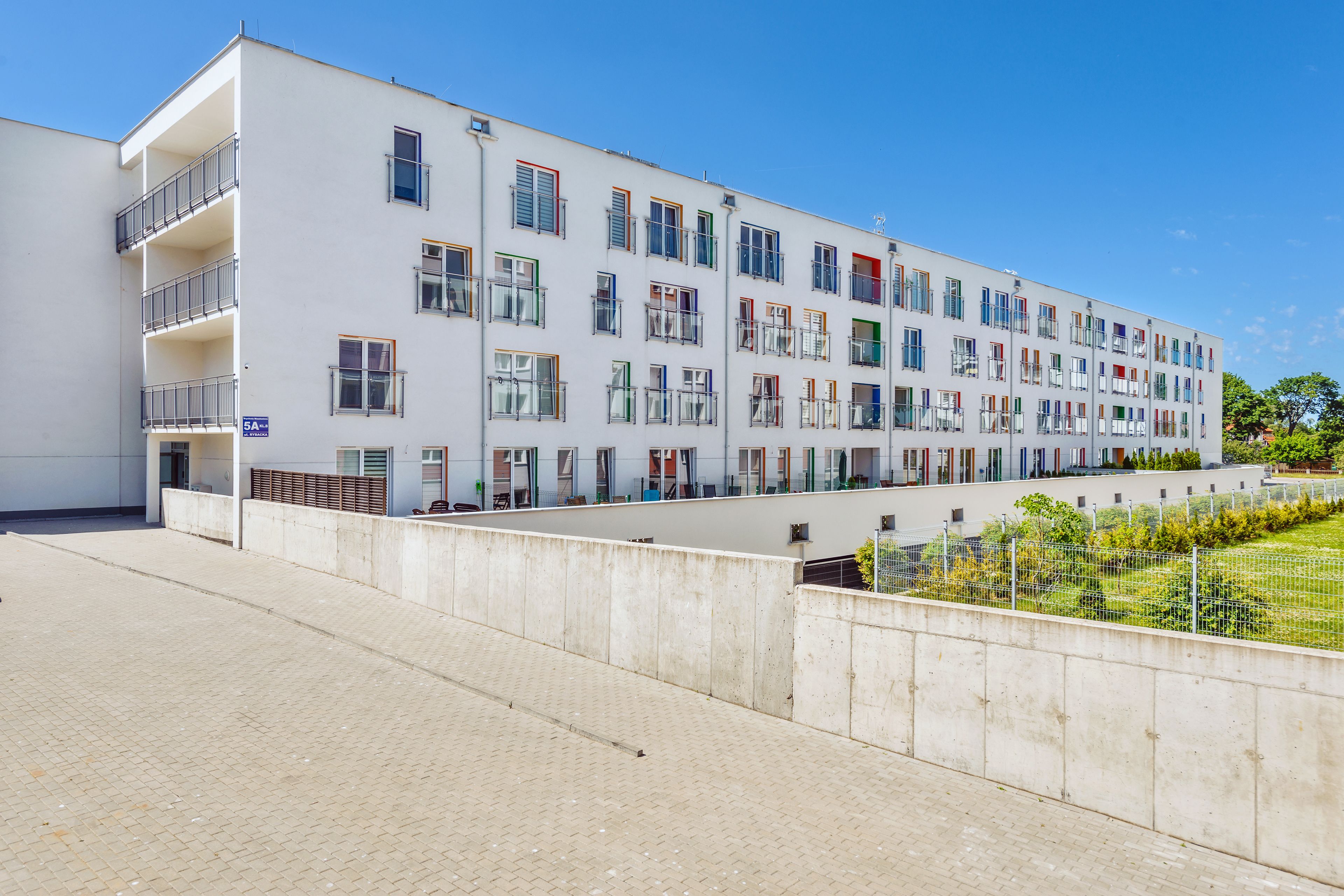 A multi-story building with balconies, a paved walkway, and a fenced garden area.