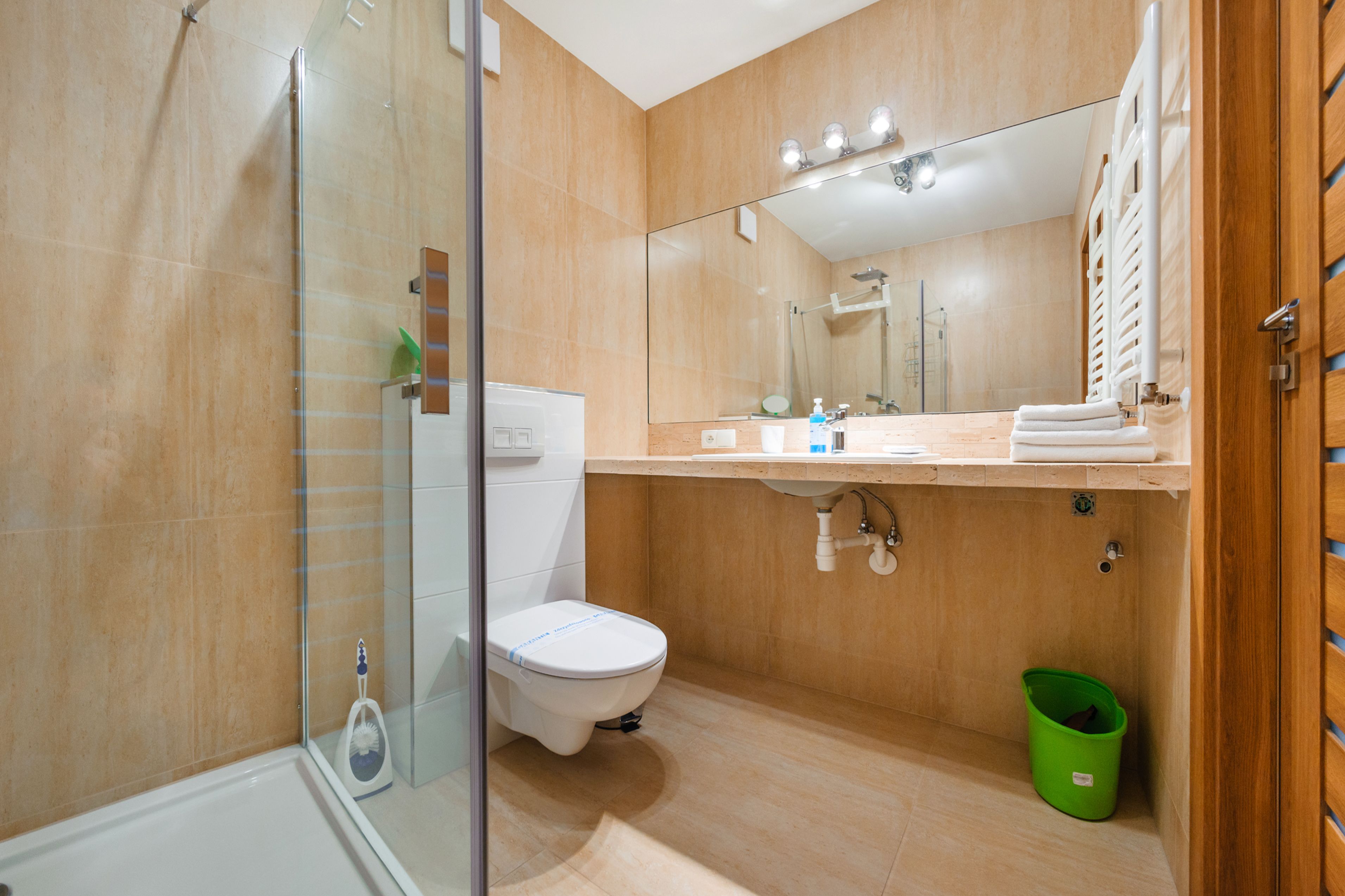 Ein modernes Badezimmer mit Duschkabine aus Glas, Toilette, Waschbecken mit Spiegel darüber und einem grünen Mülleimer.
