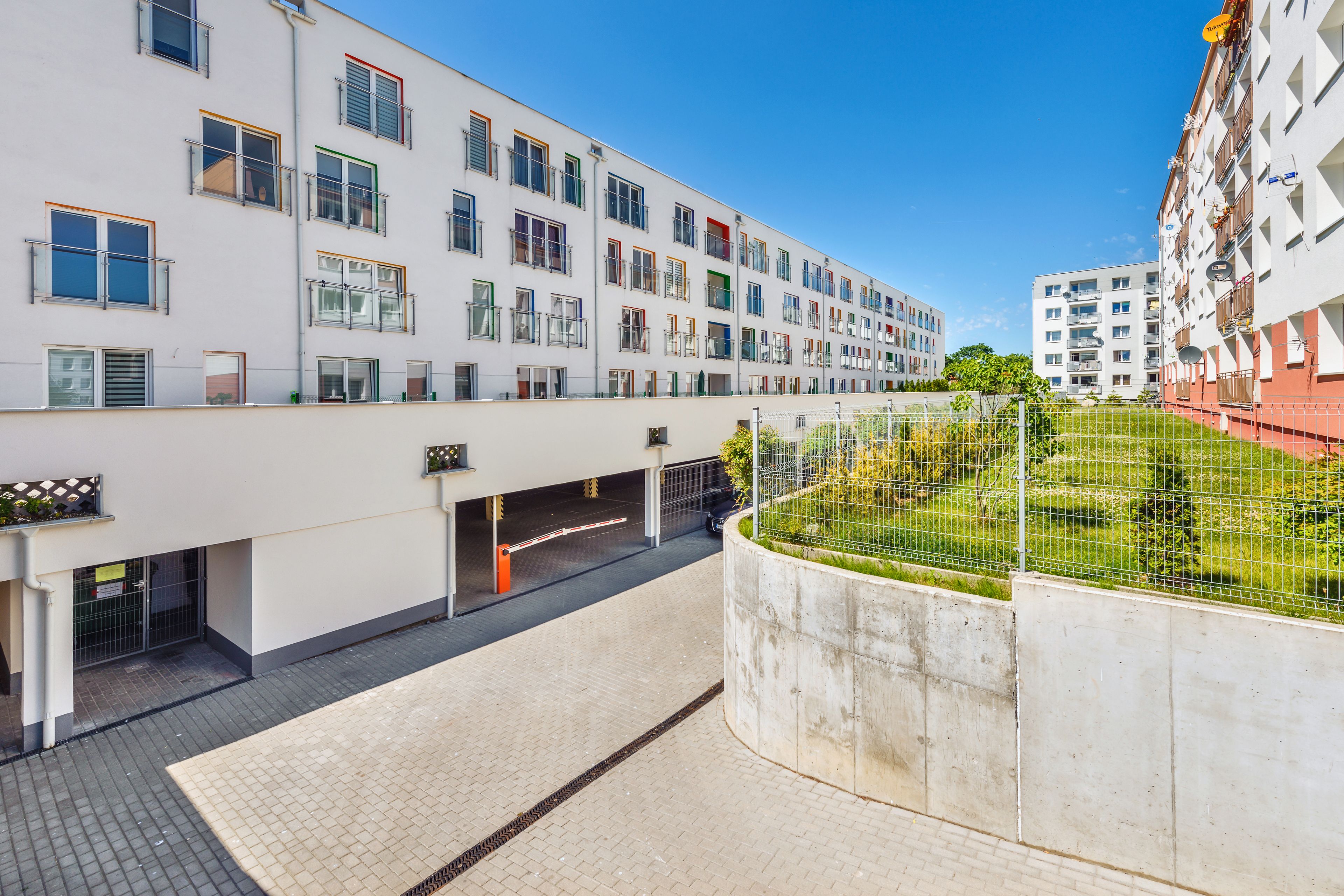 Un moderno edificio de apartamentos con varios balcones, zona de estacionamiento techada y jardín cercado.