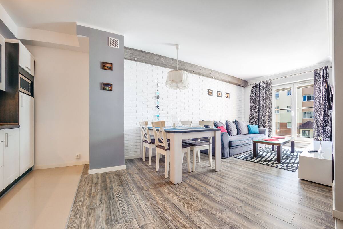 Een modern appartement met keuken, eetgedeelte en woonkamer. Het heeft een witte bakstenen muur, houten vloeren en een groot raam met kamergordijnen.