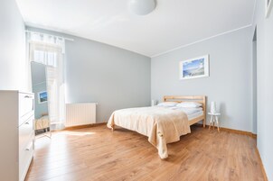 Un dormitorio con suelo de madera, una cama con cabecero, un radiador, un aparador blanco y una ventana con cortinas.