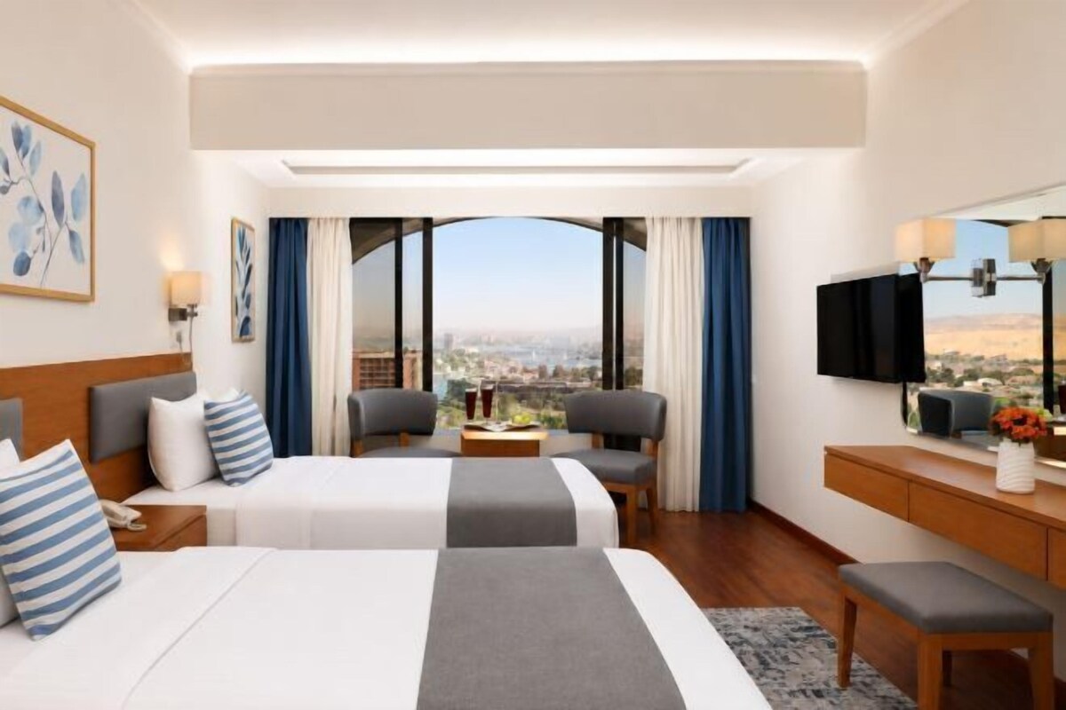 A hotel room with two beds, a desk, a TV, and a large window with a city view.