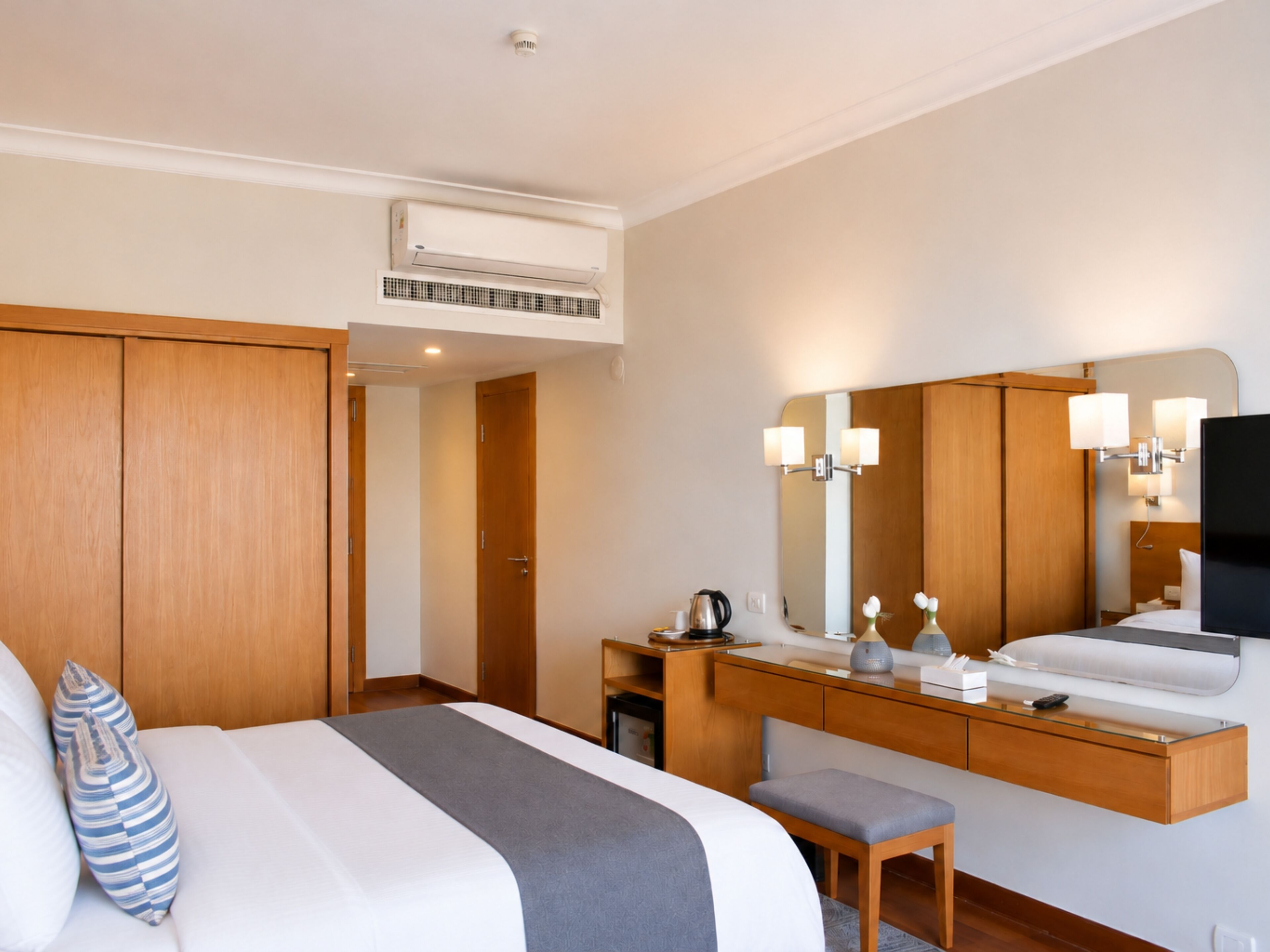 A hotel room with a bed, a desk, a chair, a mirror, and an air conditioning unit.