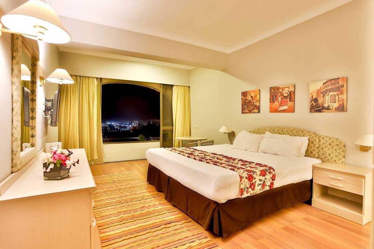 A hotel room with a large bed, a desk with a lamp, a mirror, a window with curtains, and artwork on the wall.