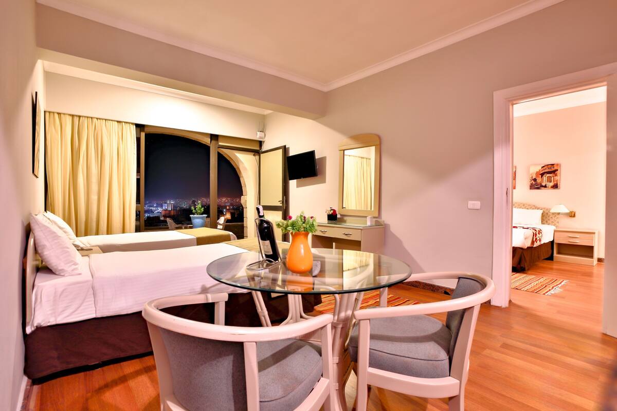 A hotel room with a bed, a glass table, two chairs, a TV, a window with curtains, and a night view outside.
