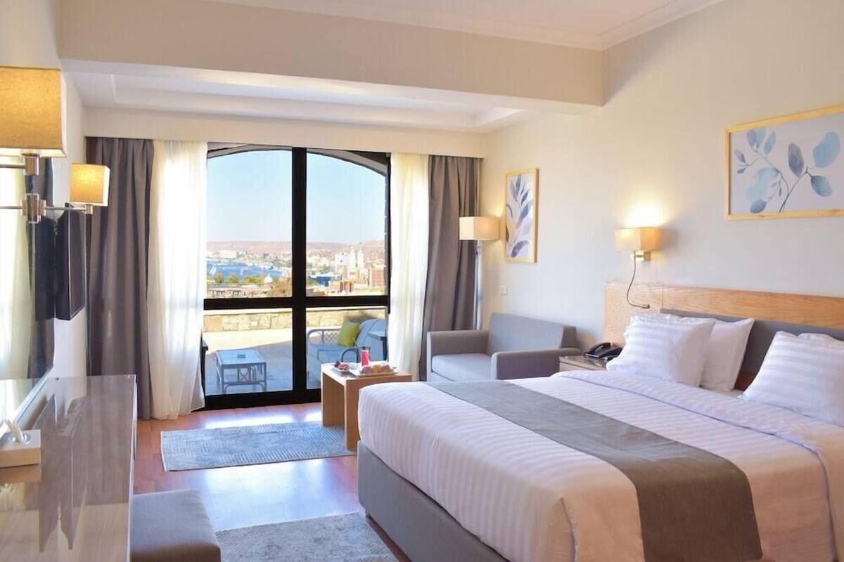 A hotel room with a large bed, a desk, a chair, a sofa, and a balcony with a city view.