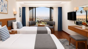 A hotel room with two beds, a desk, a chair, a TV, and a view of a cityscape. - Basma Executive Club (Aswan)