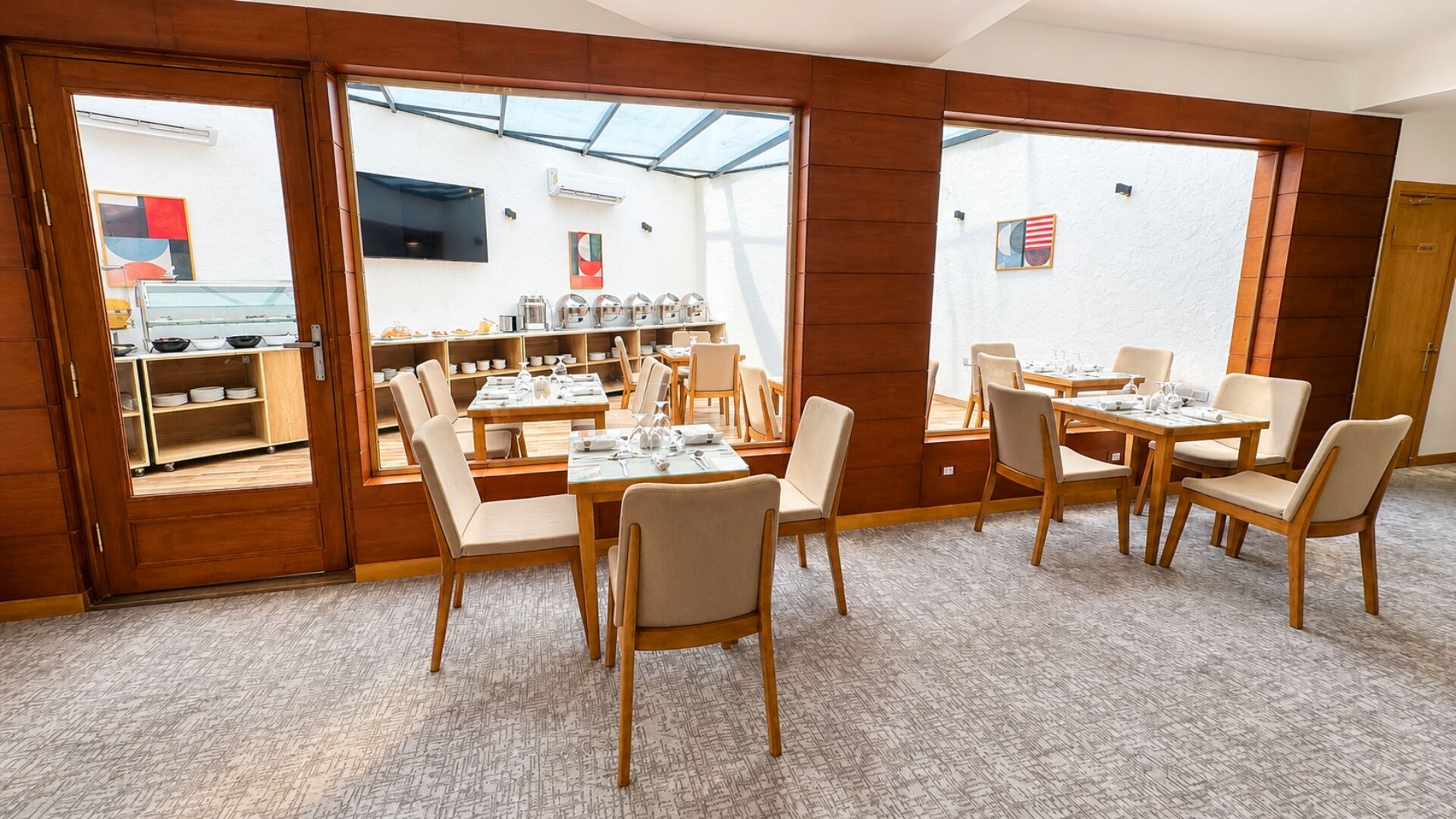 A dining area with wooden tables and chairs, a kitchen in the background, and large windows.