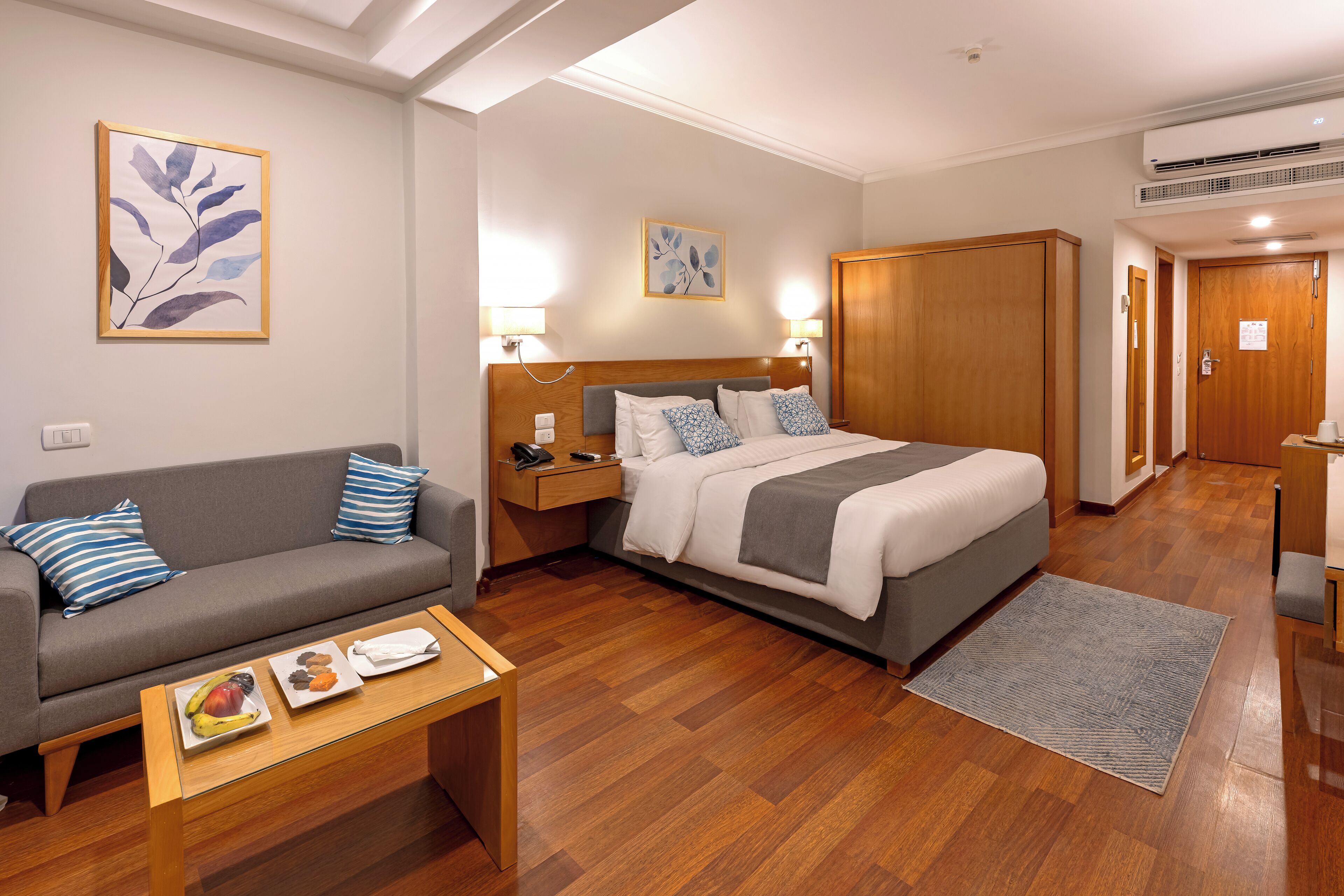 A hotel room with a bed, a sofa, a nightstand, a desk, and a small table with fruit.