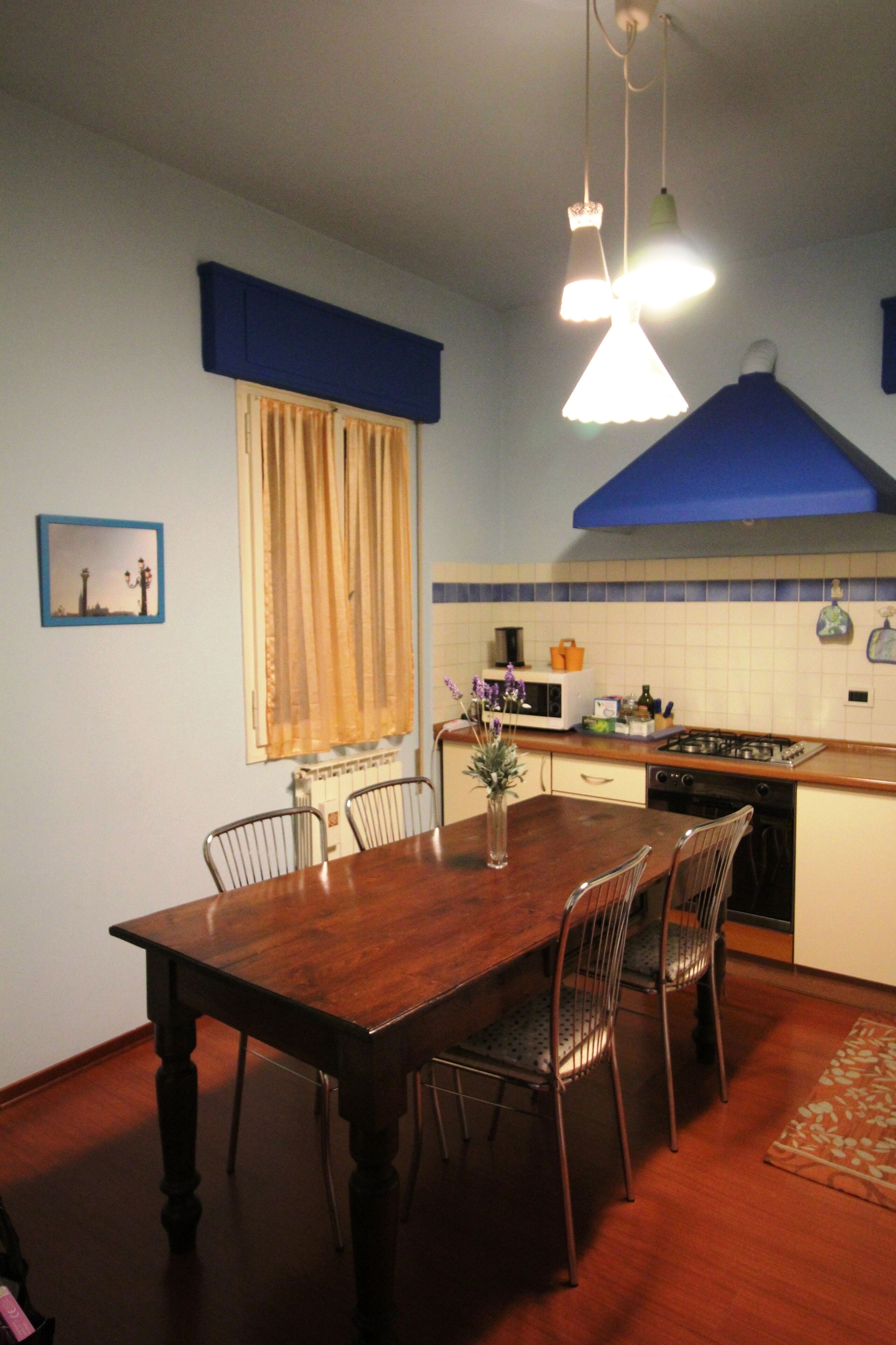 A dining area with a wooden table and chairs, a window with curtains, a kitchen with a stove and microwave, and a blue ceiling.