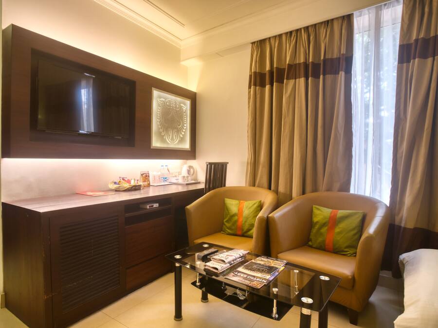 Habitación de hotel con televisión de pantalla plana sobre un escritorio de madera oscura, un sofá de cuero cómodo con cojines verdes y naranja, y una mesa de centro de vidrio con revistas.