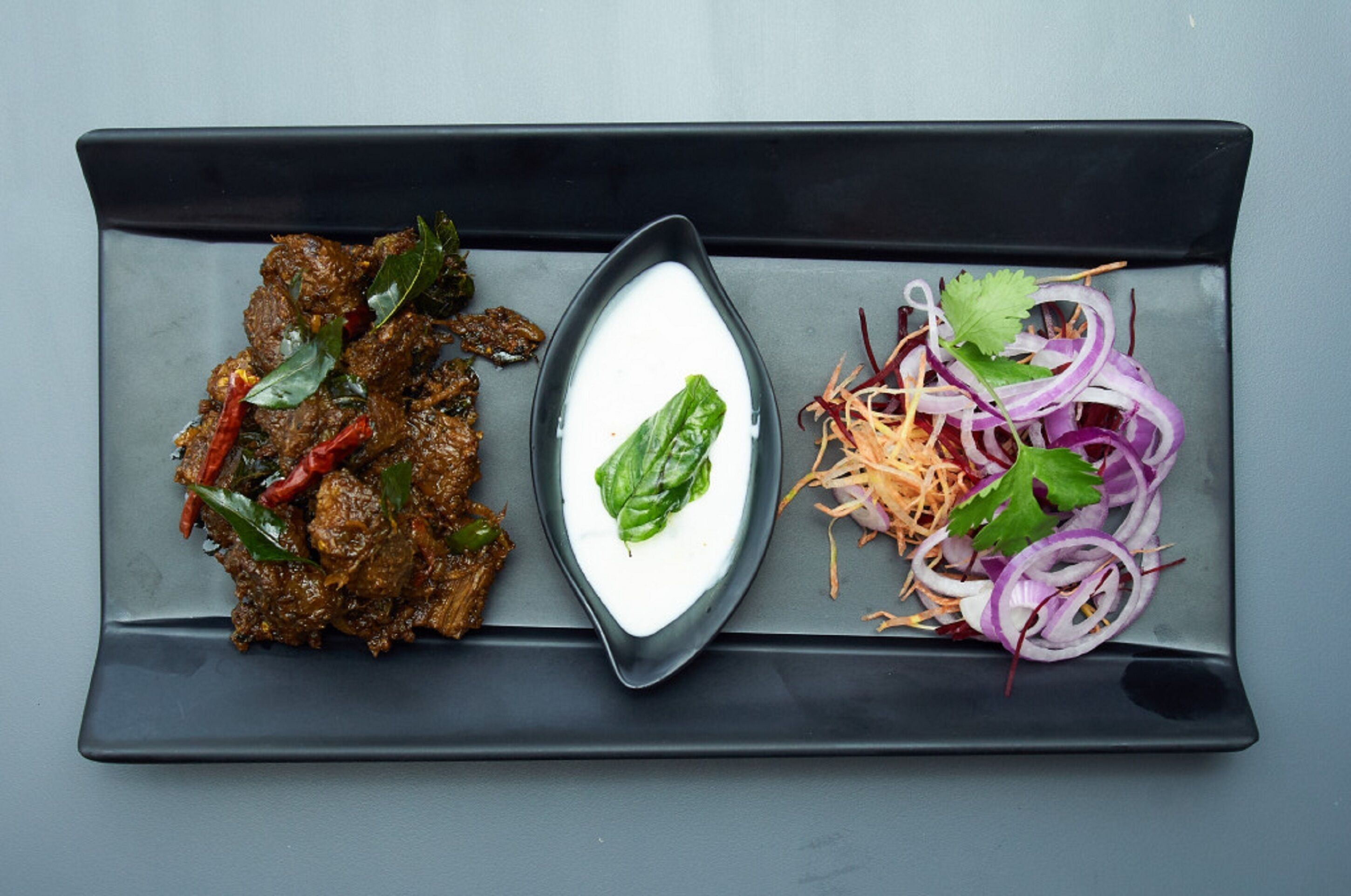 A rectangular plate with a dark sauce, a white creamy dish with a green leaf, and a side of shredded vegetables and sliced onions.