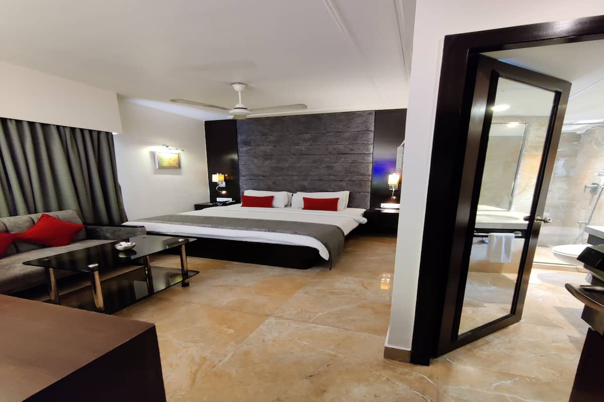 A modern hotel room with a large bed, a sofa, a coffee table, and a bathroom visible through a glass door.