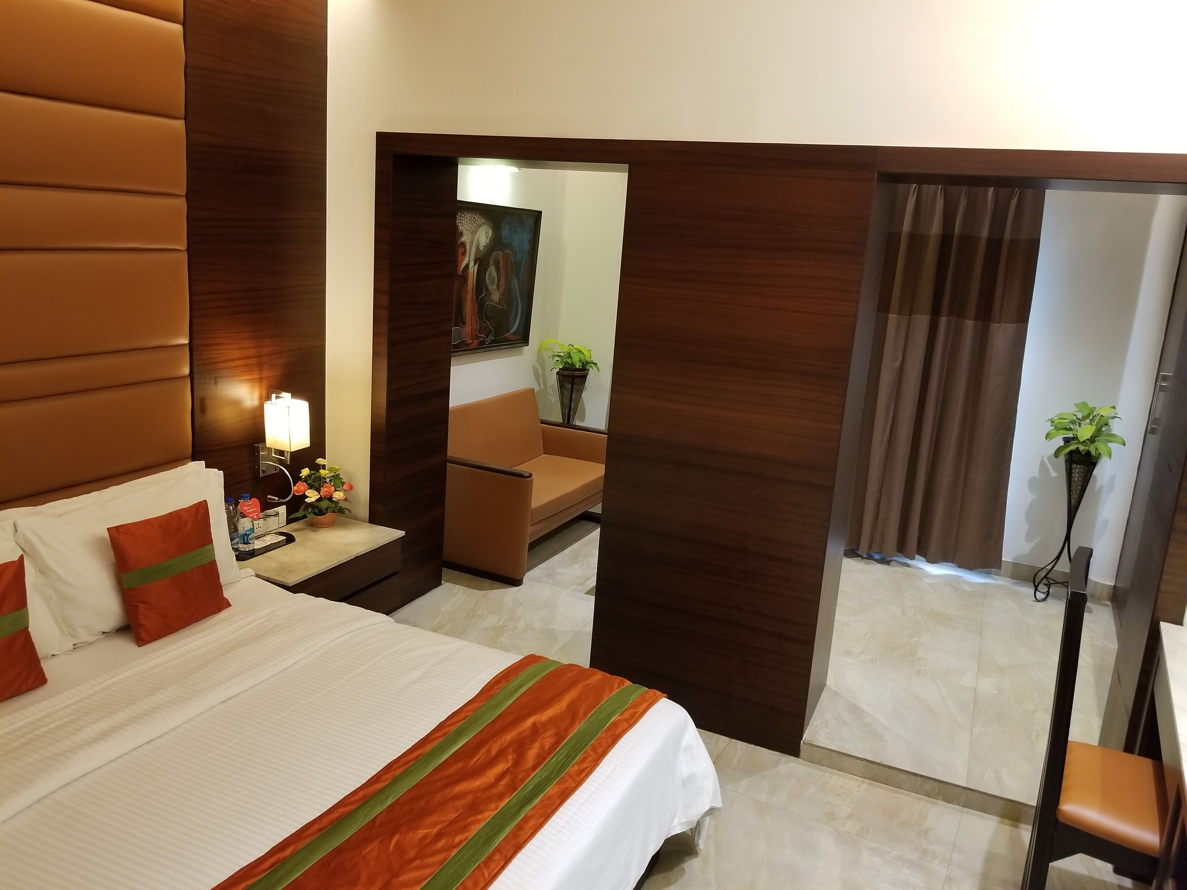 A hotel room with a large bed, a bedside table with a lamp, a small table with a vase of flowers, a sofa, and a chair.