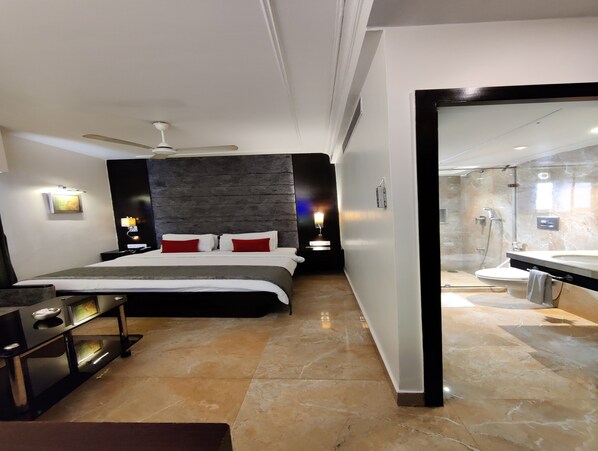 A modern hotel room with a large bed, a desk, a vanity with a mirror, and a shower area.