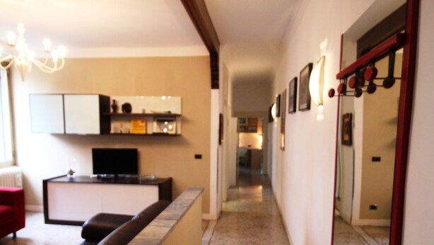 A narrow hallway with a kitchen area on the left, featuring a countertop, cabinets, and a television. A large mirror is on the right wall, reflecting part of the hallway. - Venezianamente - Casa Cavaletti (Venice)
