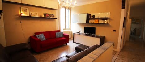 Una sala de estar con un sofá rojo, un sofá de cuero marrón, un televisor y una zona de cocina con armarios y encimera.