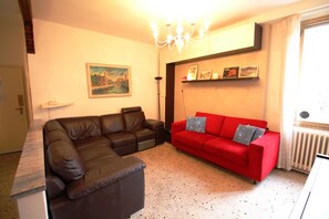 A living room with a brown leather sofa, a red sofa with pillows, a painting on the wall, and a chandelier. - Venezianamente - Casa Cavaletti (Venice)