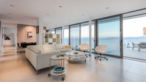 A modern living room with a white sofa, two white chairs, a coffee table, and a bookshelf. Large windows offer a view of the sea. - 600m² homm Luxury Villa Sea Side Evia 16ppl (Istiaia-Aidipsos)