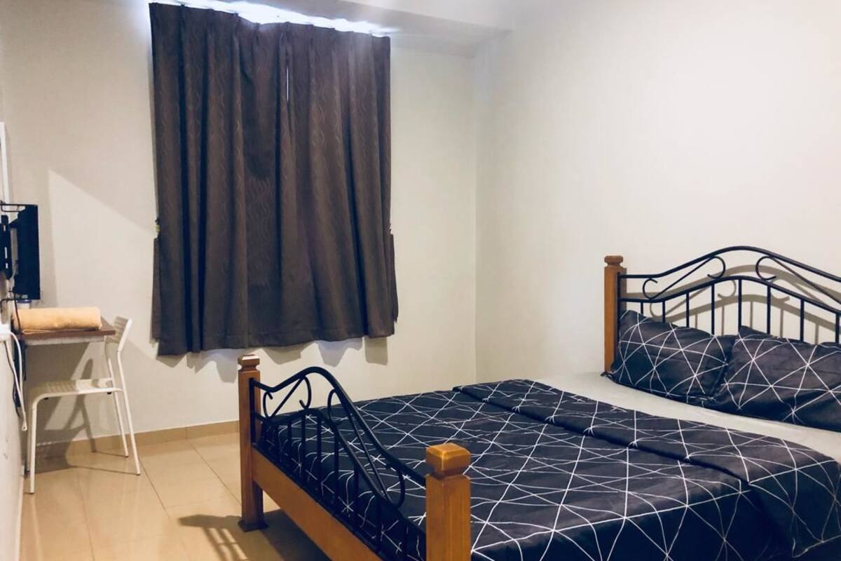 A single bed with a wooden frame and black metal headboard, a small white desk with a chair, and a window with dark curtains.