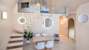 Un interior moderno con una escalera que lleva a un altillo, una zona de comedor con una mesa de madera y sillas blancas, y una lámpara colgante.
