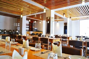 A restaurant with wooden ceiling panels, multiple dining tables with white napkins, and a bar area in the background.