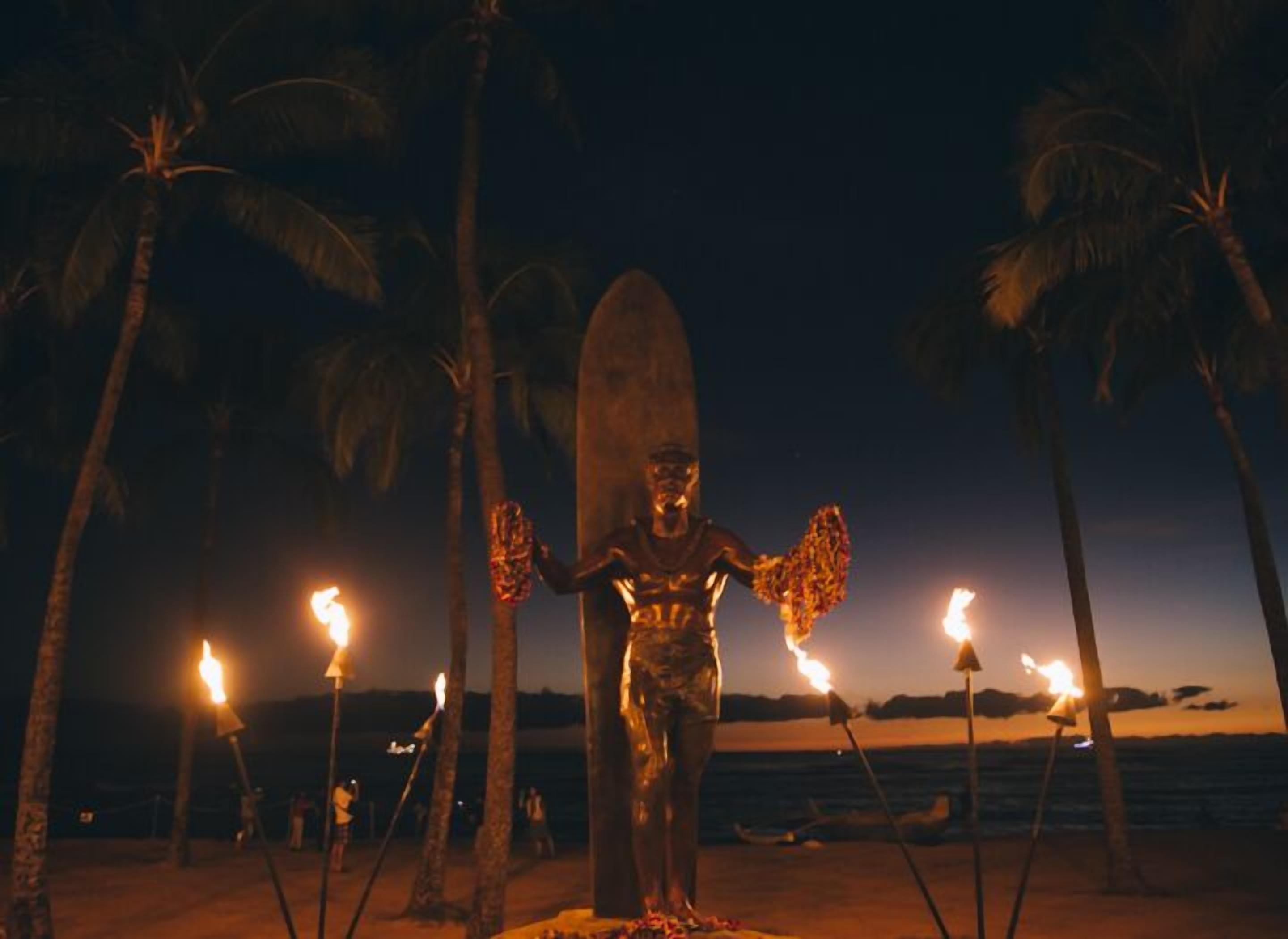 Eine Statue mit einem Surfbrett, umgeben von Fackeln an einem Strand bei Dämmerung.
