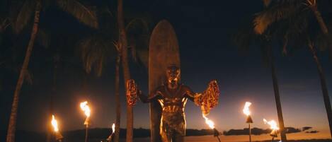 Eine Statue mit einem Surfbrett, umgeben von Fackeln an einem Strand bei Dämmerung.