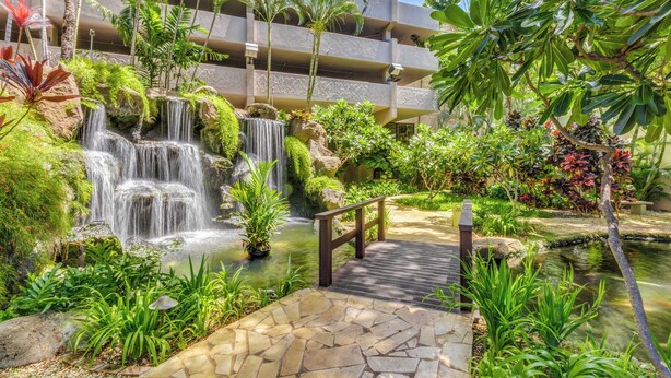 A lush garden with a waterfall, a stone pathway, and a view of a multi-story building with balconies.