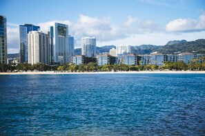 Un paisaje urbano costero con edificios modernos altos, una playa y montañas al fondo.