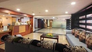 A hotel lobby with a reception desk, a sofa with patterned cushions, a coffee table with flowers, and a large window with patterned curtains. - Pushpak Grande (Kondotti)