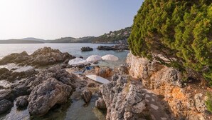 Uma praia rochosa com guarda-sóis e espreguiçadeiras, vista clara do mar e um morro coberto de árvores.