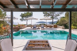 Un espace aménagé au bord de la piscine, avec des chaises longues et des parasols, un foyer extérieur et une vue sur la mer.