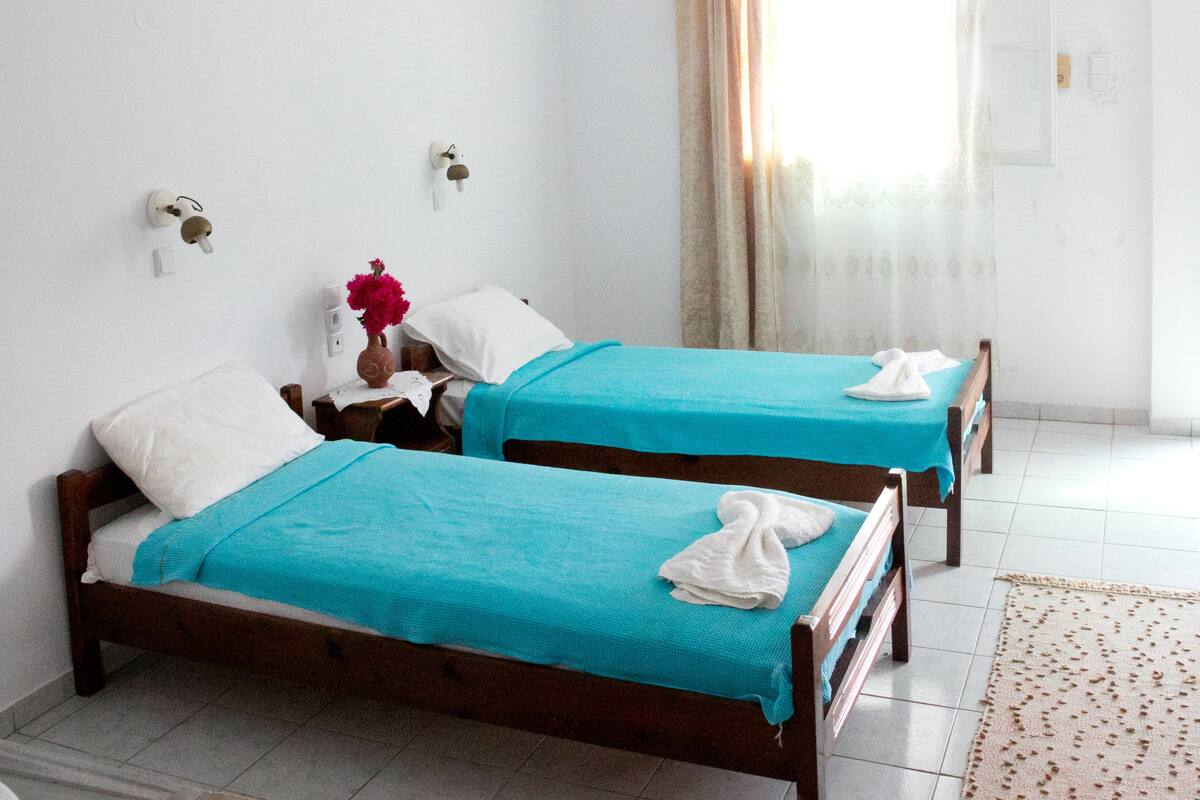 A room with two beds, a ceiling fan, a window with curtains, and a small table.