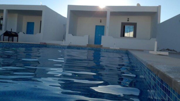 A swimming pool with two white buildings in the background, each with a blue door. - Dar Aesha (Aghir)