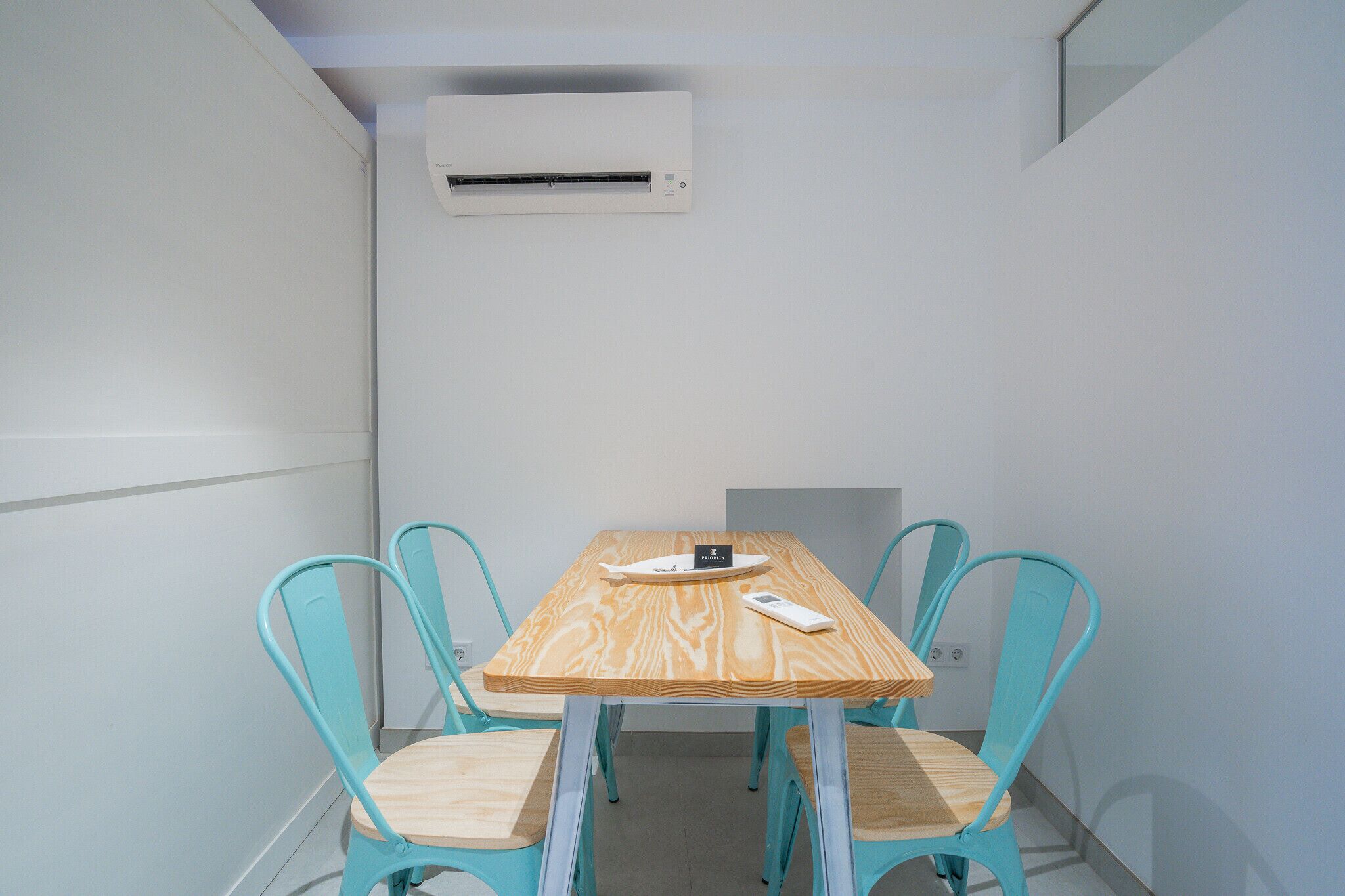 Quarto pequeno e moderno com uma mesa de madeira e quatro cadeiras azul-turquesa, um aparelho de ar condicionado na parede e uma pequena janela.
