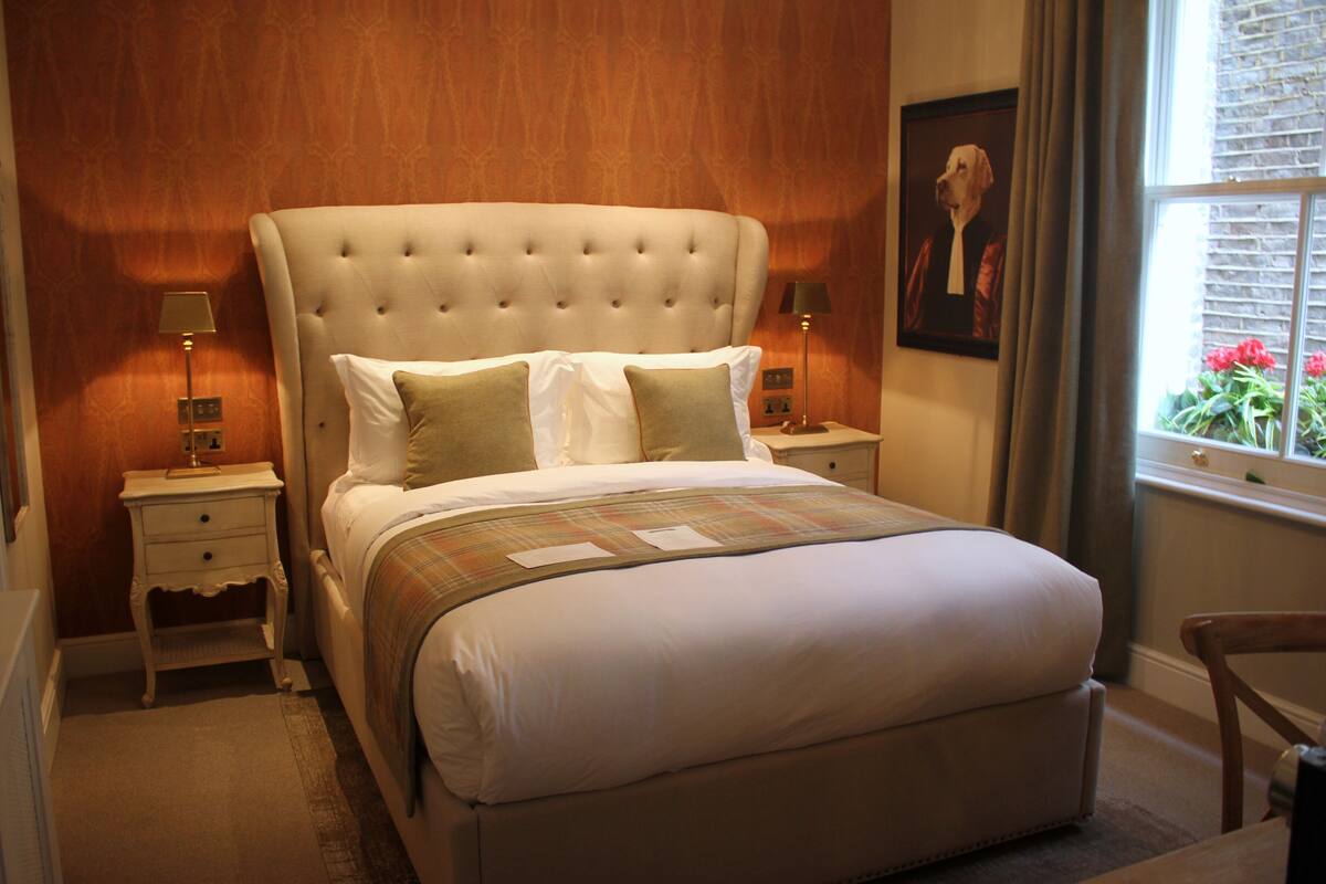 A bedroom with a tufted headboard, a bed with white linens, two bedside tables with lamps, a framed picture on the wall, and a window with a view of flowers.