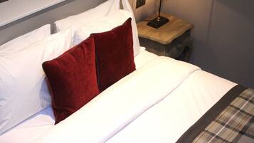 A neatly made bed with white linens and a red pillow, a small wooden nightstand with a lamp, and a plaid blanket.