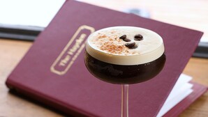 A dark-colored beverage with a frothy top and chocolate chips on a glass, placed on a surface with a maroon book in the background. - The Hayden Pub & Rooms (London)