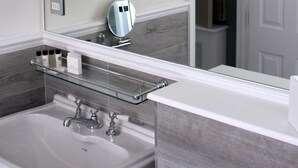 A bathroom with a large mirror, two wall-mounted lights, a glass shelf with toiletries, and a sink with a faucet. - The Hayden Pub & Rooms (London)