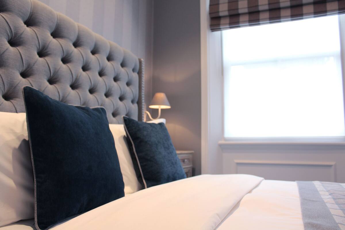 A bed with a tufted headboard, two pillows, a bedside lamp, and a window with blinds.
