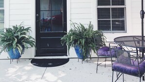 Un bâtiment blanc avec une porte d’entrée noire, deux plantes en pot, un parapluie rouge et du mobilier d’extérieur.