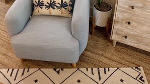 Un salon avec un fauteuil bleu clair, une petite table d’appoint en bois, un meuble blanc et une fenêtre avec des stores.