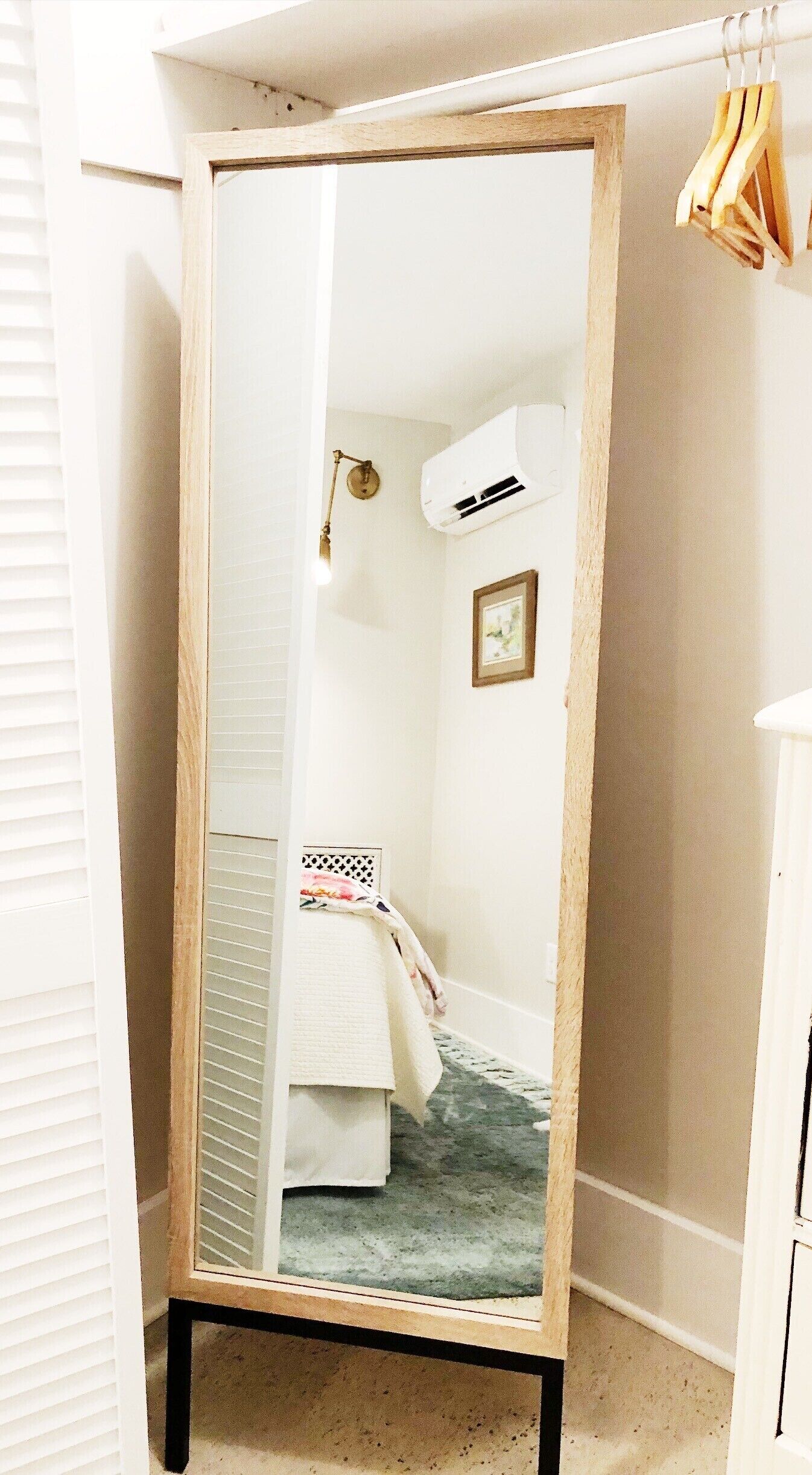 A room with a wooden-framed mirror, a bed with a floral bedspread, a wall-mounted air conditioner, a small framed picture, and a hanging clothes rack.